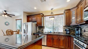 A modern kitchen with wooden cabinets, a stainless steel refrigerator, a built-in oven, and a large island with a granite countertop. - Beautiful home with private pool, outdoor kitchen & enclosed yard (Indio)
