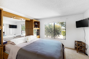 Ein Schlafzimmer mit einem großen Bett, einem Holz-Kopfteil, einem an der Wand montierten Fernseher und einem Fenster mit Blick auf Bäume.