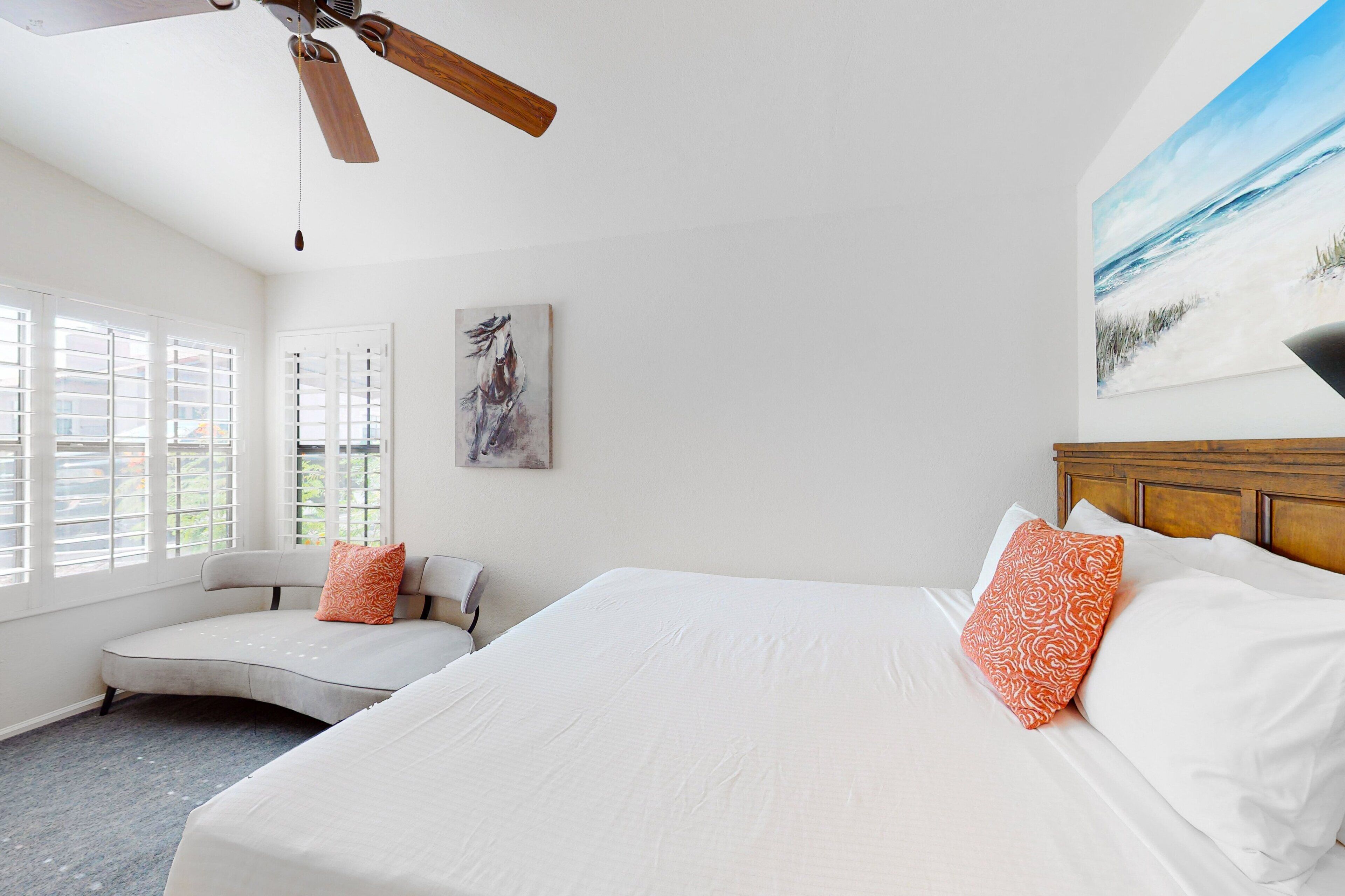 A bedroom with a bed, a curved bench, a ceiling fan, a window with blinds, and a painting on the wall.
