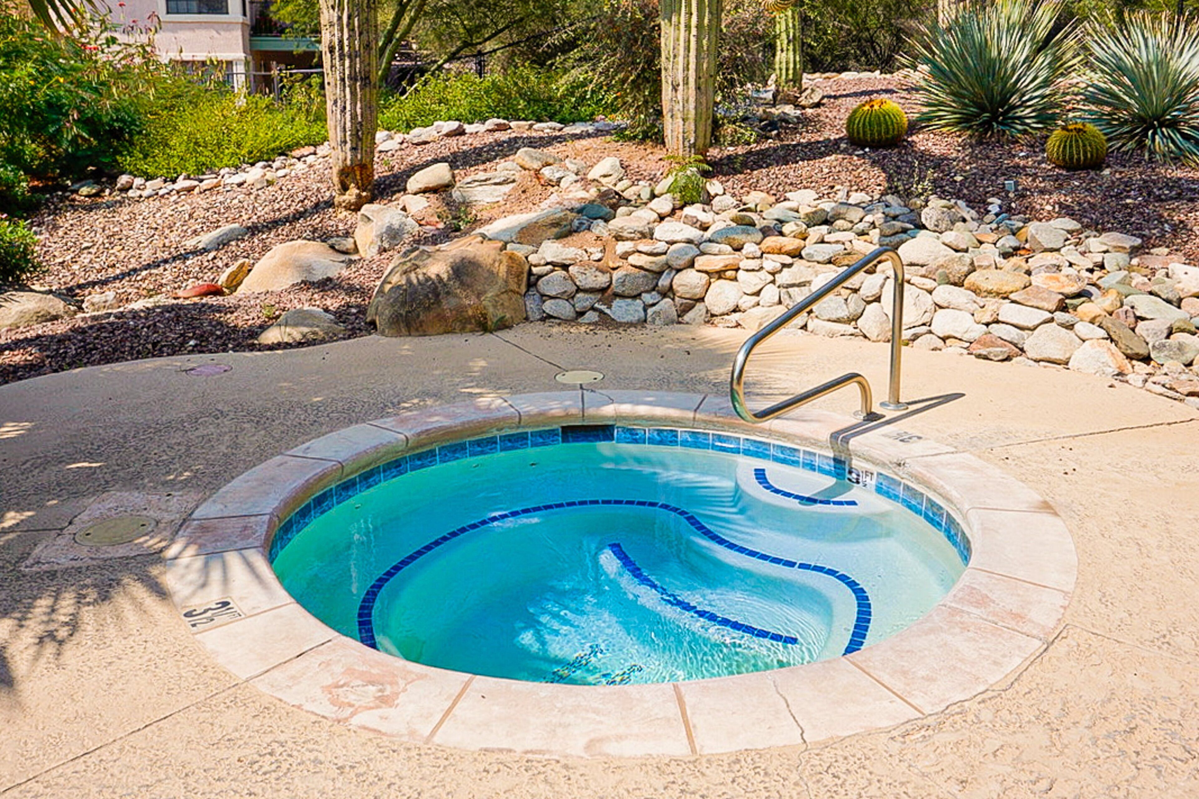 Una piscina circular con borde de piedra, rodeada por un jardín ajardinado con rocas y cactus.