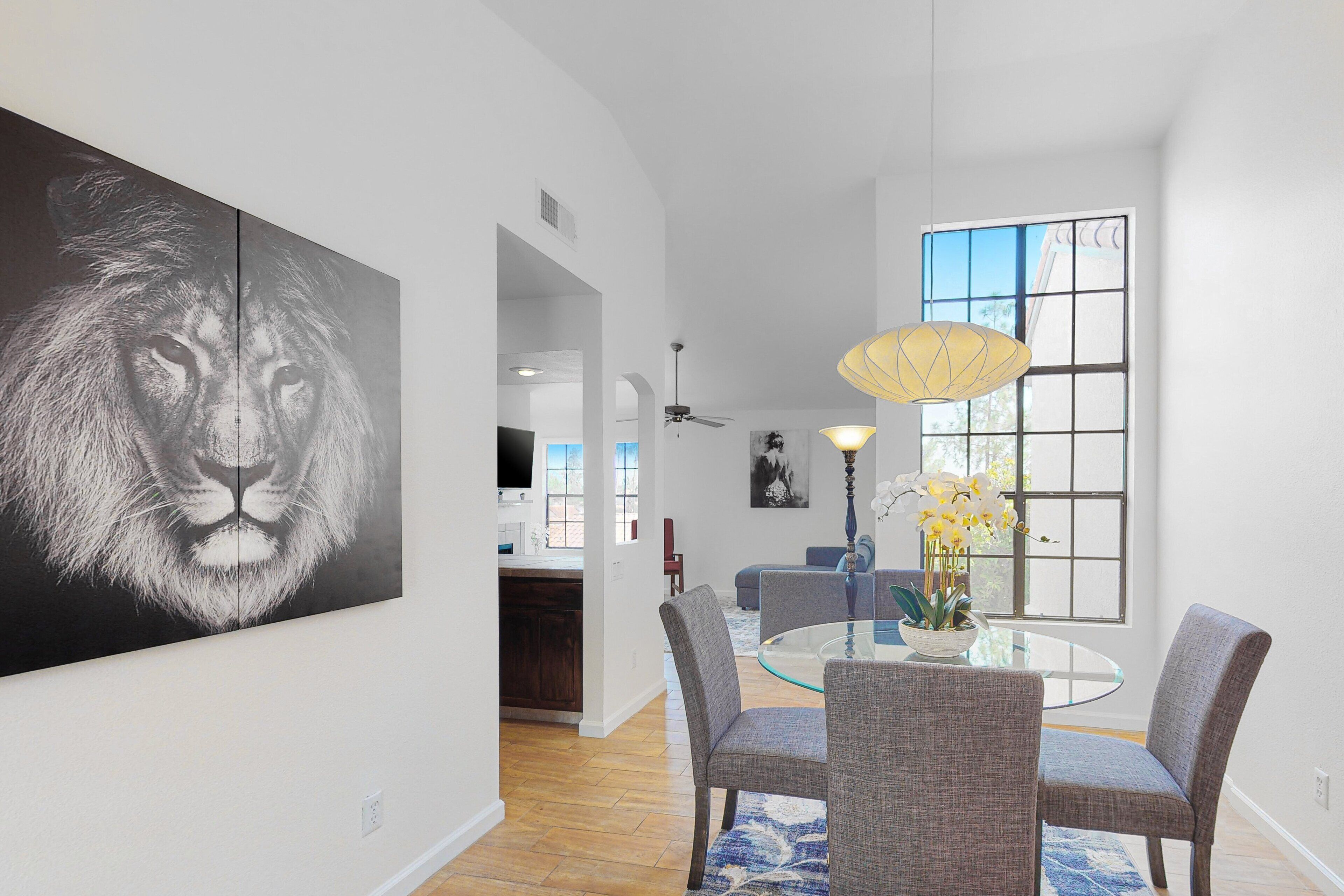 Un coin repas avec une table en verre, quatre chaises, une grande œuvre d’art représentant le visage d’un lion et une fenêtre donnant sur un beau paysage.
