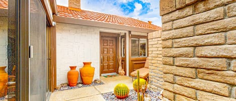 A courtyard with a white brick wall, a wooden door, and a tiled roof. There are three cacti and two large vases on the ground.