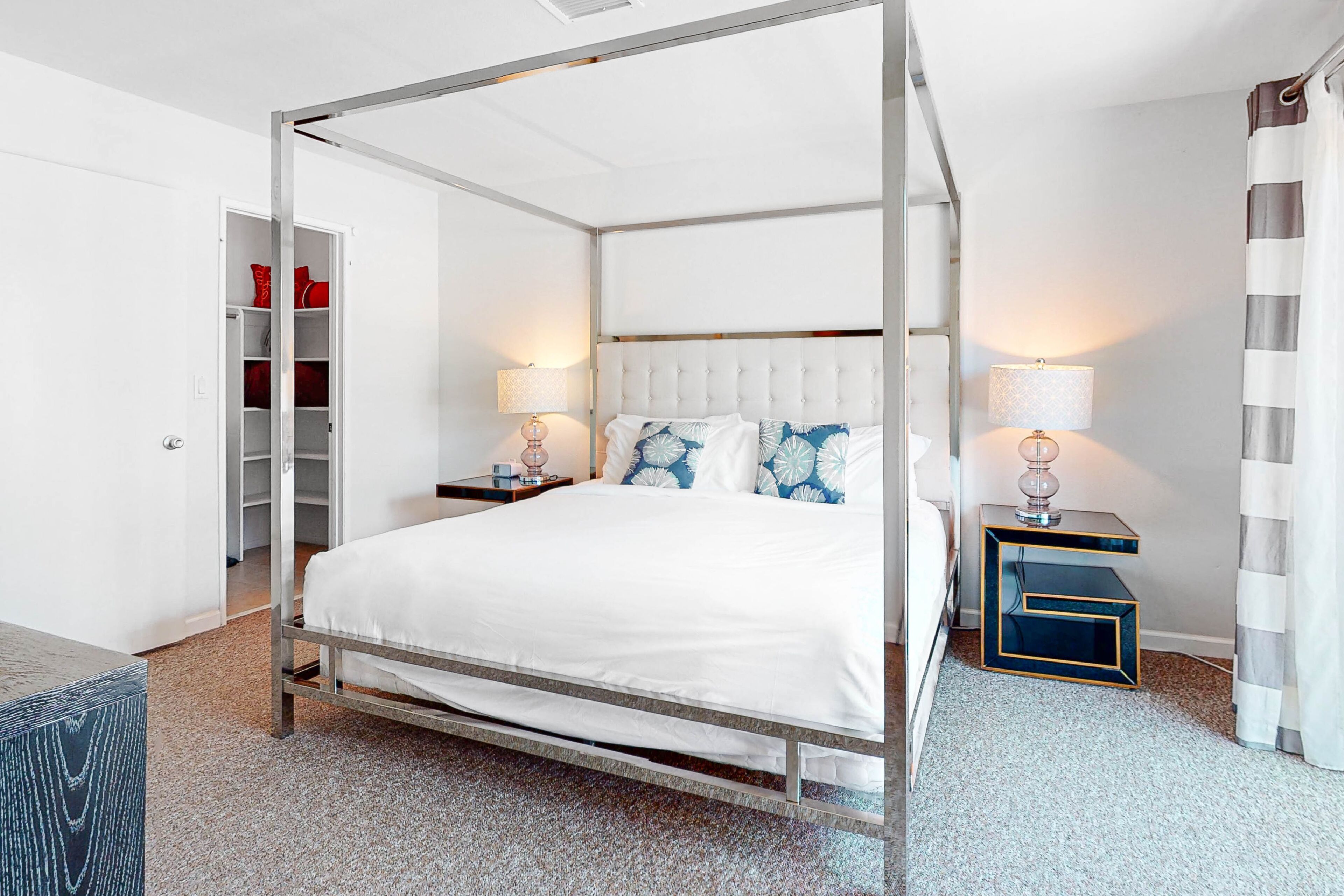 Een hemelbed met witte beddengoed, twee nachtkastjes met lampen, een garderobe en een gestreept gordijn.