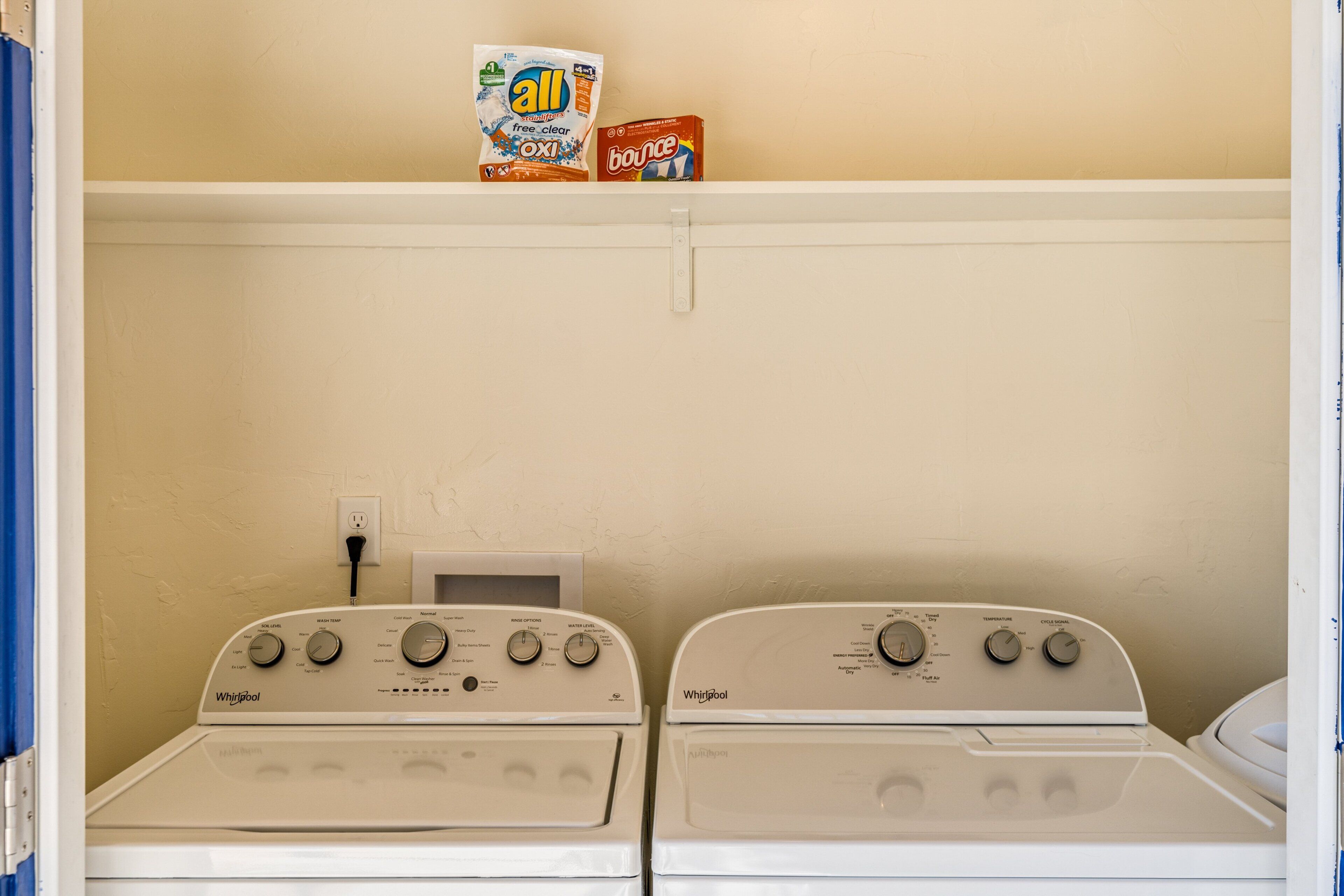 Uma lavanderia com duas máquinas de lavar e secar Whirlpool, uma prateleira com caixas de detergente e uma tomada.