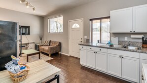 Una cocina moderna con armarios blancos, encimera de granito y horno empotrado. Hay un comedor con mesa y sillas, y una sala de estar con sofá y televisor.