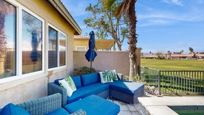 Eine Terrasse mit einem blauen Ecksofa, einem Sonnenschirm und Blick auf einen gepflegten Rasen sowie entfernte Gebäude.