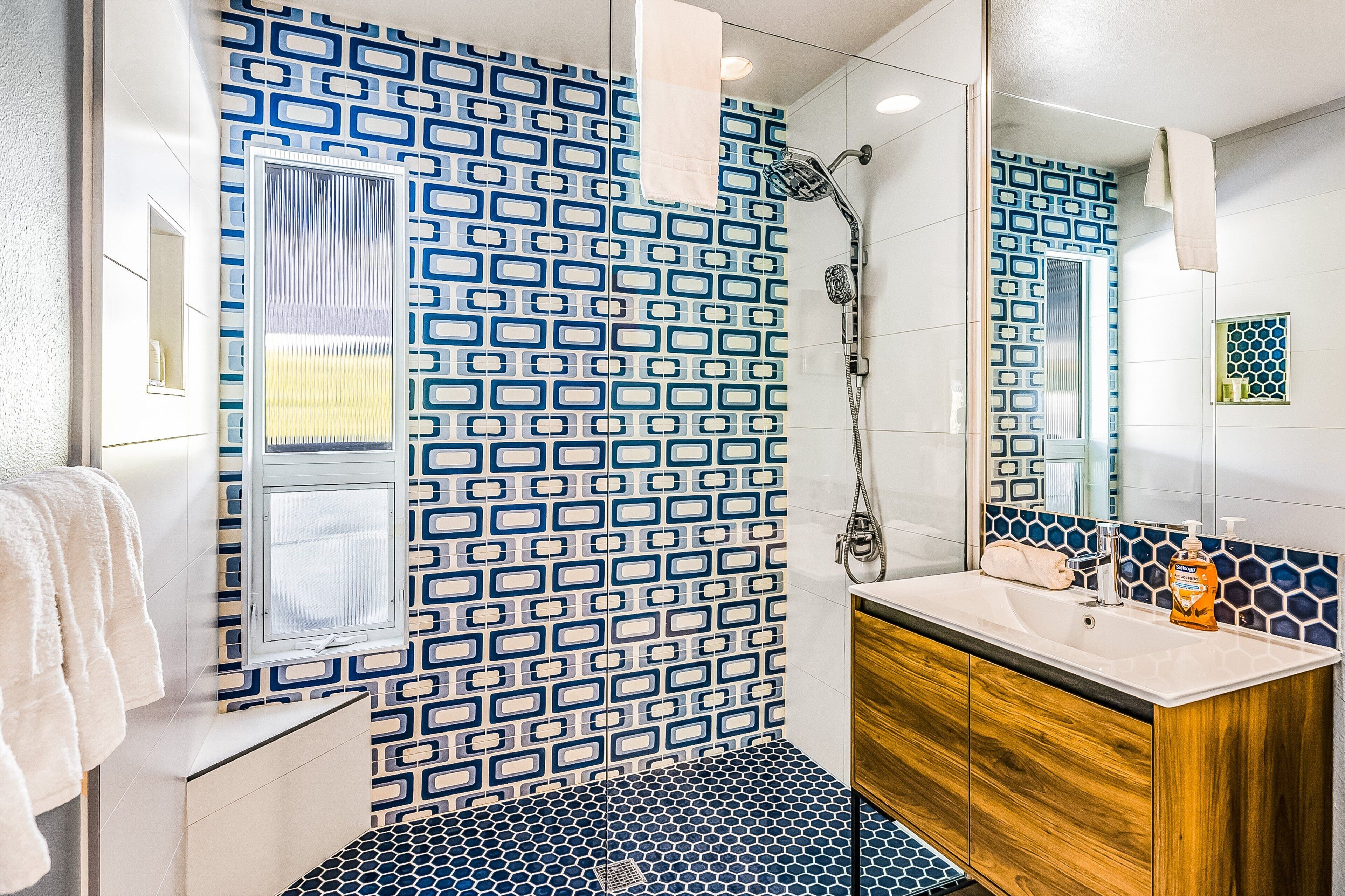 A bathroom with a patterned tile wall, a shower, a sink with a wooden vanity, and a mirror.