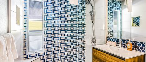 A bathroom with a patterned tile wall, a shower, a sink with a wooden vanity, and a mirror.