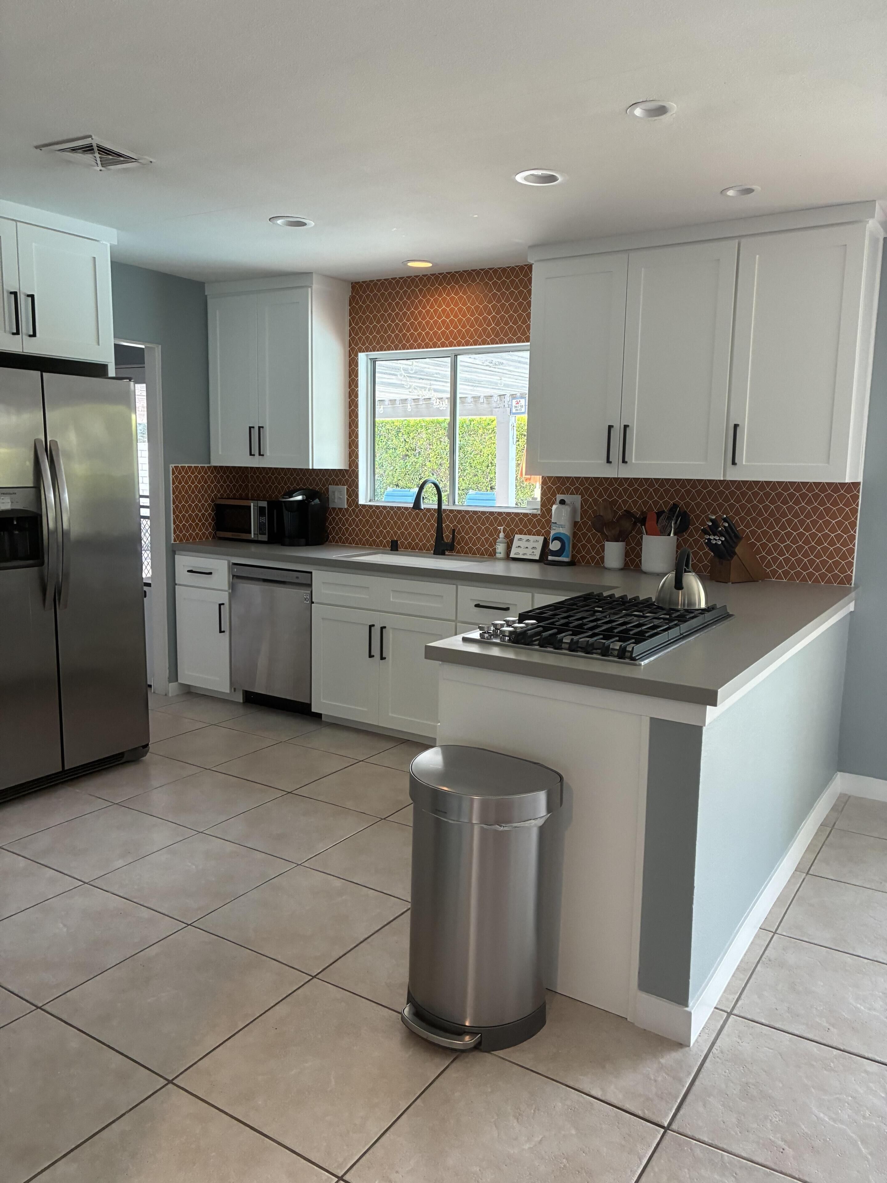 A modern kitchen with white cabinets, stainless steel appliances, a window with a view, and a tiled floor.