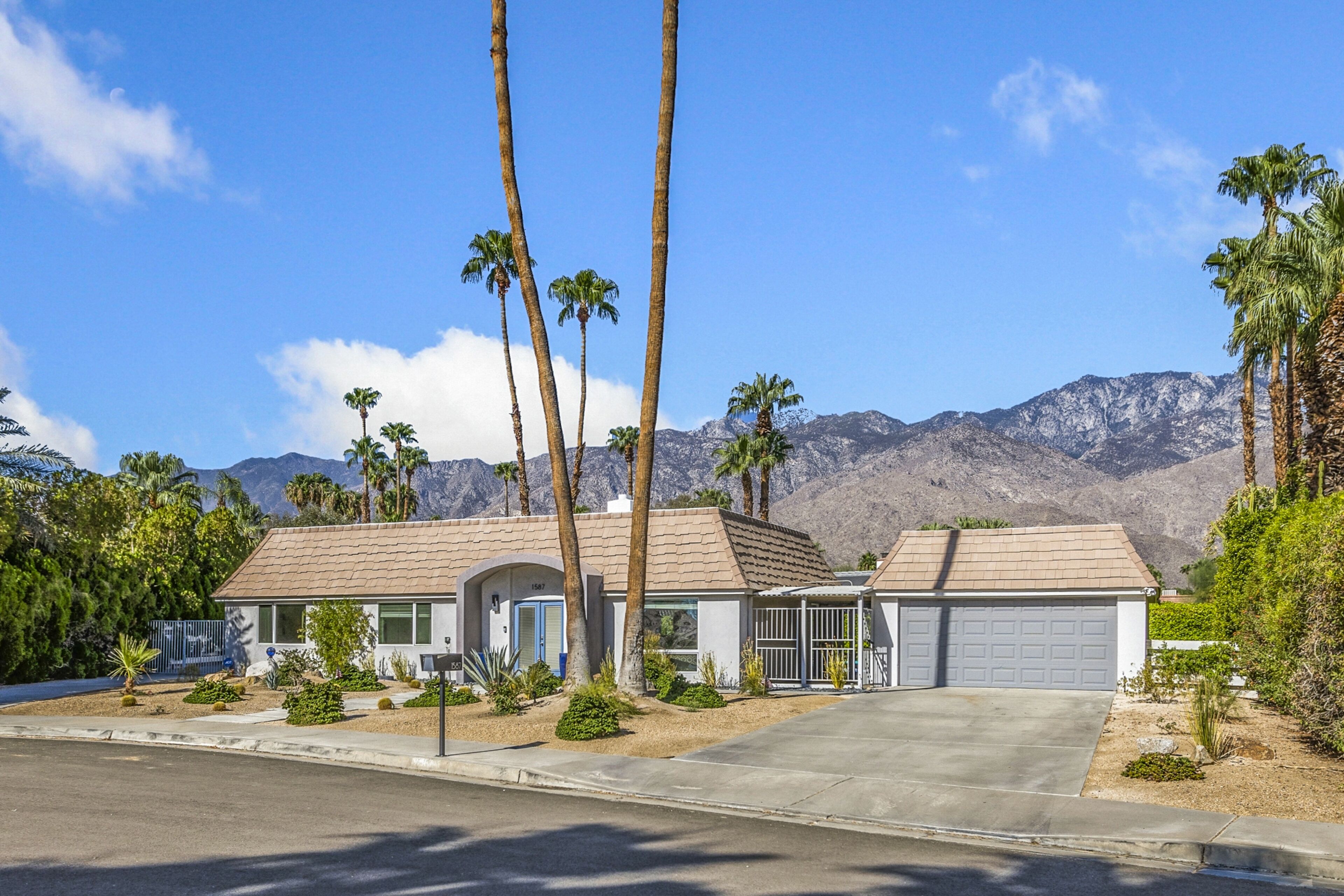 A single-story house with a garage, surrounded by palm trees and mountains in the background.
