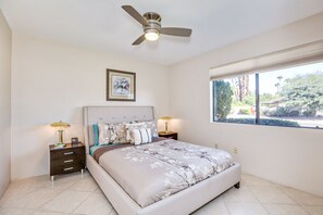A bedroom with a bed, two bedside tables, a ceiling fan, a window with a view of greenery, and a framed picture on the wall. - Pleasant home with private pool, spa & private gas grill - close to downtown (Palm Springs)