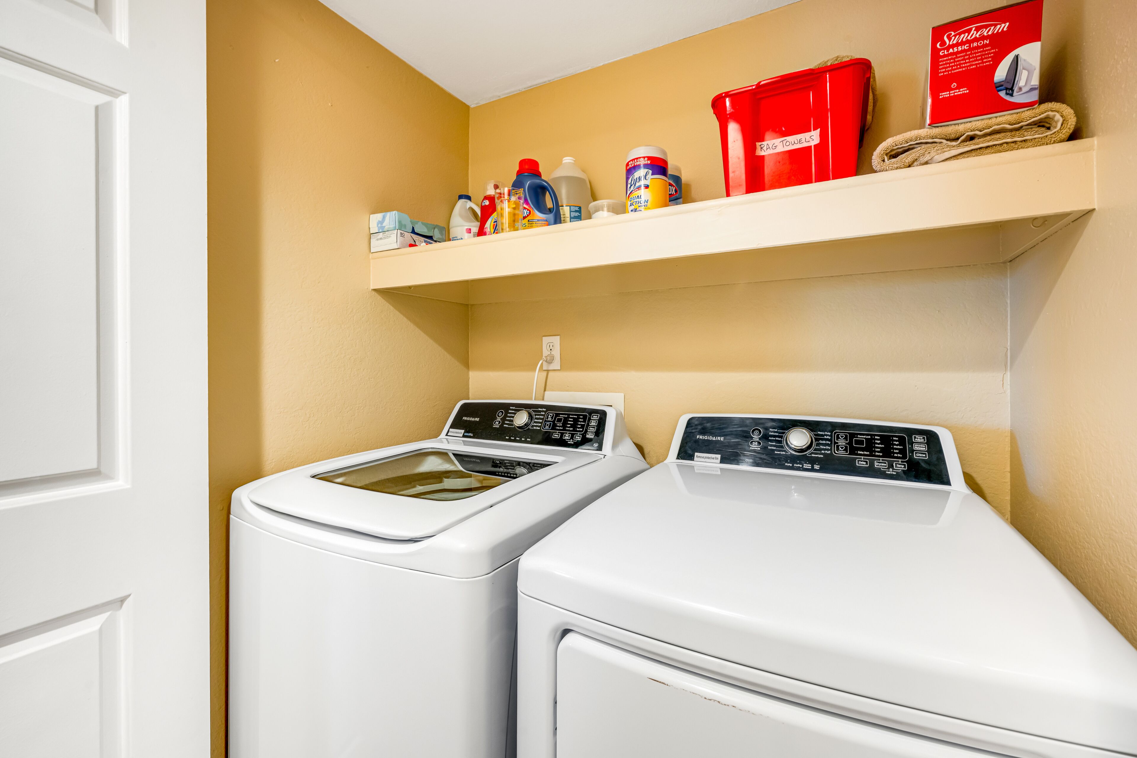 A laundry area with two white front-loading washers, a shelf with cleaning supplies, and a red laundry basket.