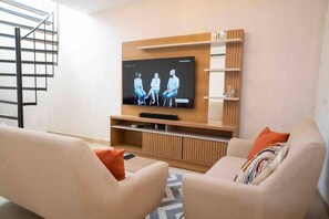 A modern living room with a flat-screen TV mounted on a wooden wall unit, two beige sofas with colorful cushions, and a staircase to the left. - Casa Moneda 3 House Old city Pool near clock tower (Cartagena de Indias)