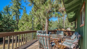 A wooden deck with a glass table and striped chairs, surrounded by tall pine trees.
