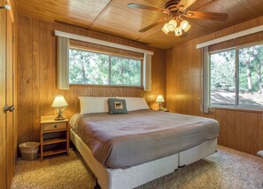Dormitorio con paneles de madera, una cama, dos lámparas de noche, un ventilador de techo y ventanas amplias.