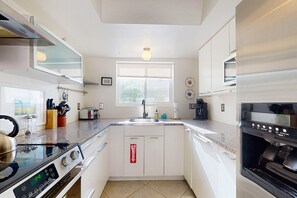 A modern kitchen with a stove, sink, and oven. There is a window with blinds, a countertop with various items, and a fire extinguisher on the wall. - Recently Updated Home w/ Private Patios: 15 Mins From Downtown Palm Springs (Desert Hot Springs)