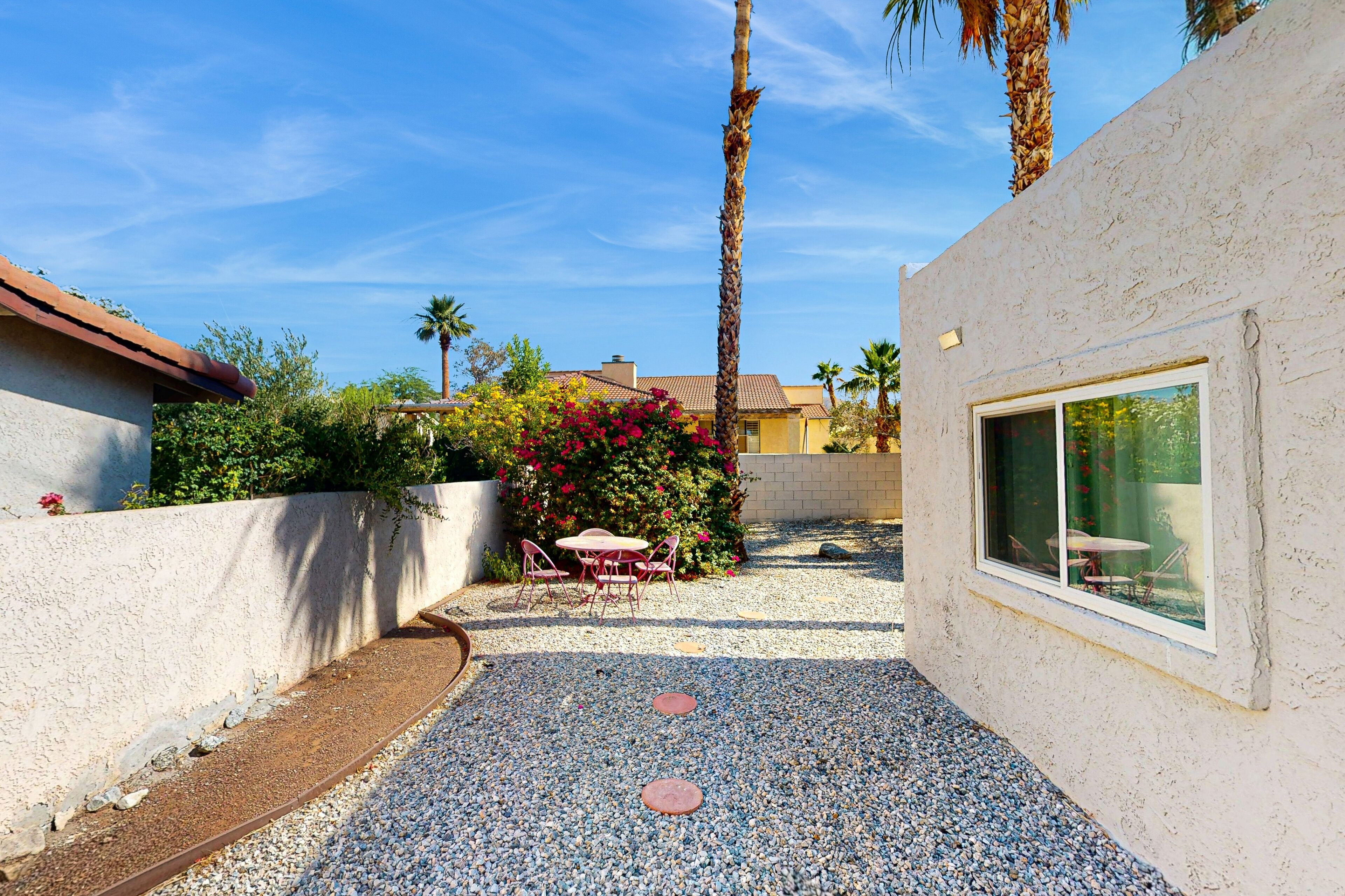 A patio area with a table and chairs, surrounded by a gravel path and palm trees.