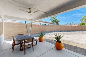 Un patio exterior con una mesa y sillas, dos plantas en macetas y vistas a una piscina y casas al fondo.