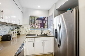 A modern kitchen with white cabinets, a stainless steel refrigerator, a built-in oven, and a window with blinds. - Beautifully updated mid-century condo with shared pool/spa & mountain views (Palm Springs)