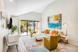 A modern living room with a white sofa, yellow armchairs, and a coffee table. There is a large abstract painting on the wall, a flat-screen TV mounted on the wall, and a floor lamp. - Beautifully updated mid-century condo with shared pool/spa & mountain views (Palm Springs)
