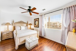 Ein Schlafzimmer mit Holzboden, einem Bett mit Kopfteil, einem Deckenventilator, einem Nachttisch mit Lampe und einem Fenster mit Jalousien.