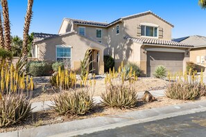A two-story house with a garage, surrounded by desert plants and palm trees. - Inviting and unique dog-friendly home with pool, pool spa, firepit, & W/D (Indio)