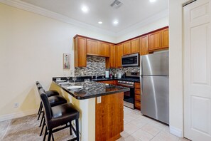A kitchen with wooden cabinets, a stainless steel refrigerator, a microwave, and a granite countertop island with bar stools. - Clean and welcoming king-suite w/ shared pool & hot tub (La Quinta)