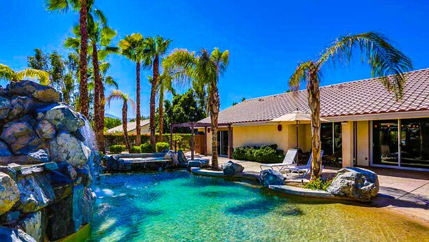 A clear pool with rocks, palm trees, and a building with a tiled roof. - Luxurious Dog-Friendly home with an outdoor kitchen, private pool, spa, & casita (Bermuda Dunes)