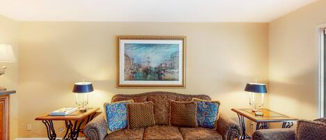 A living room with a sofa, two side tables with lamps, a framed painting, and a coffee table with magazines.