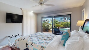 Ein Schlafzimmer mit einem Bett, einem Deckenventilator, einem Fernseher und einer Glasschiebetür, die auf einen Balkon mit Gartenmöbeln führt.