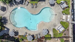 En vy från ovan av en swimmingpool med en vågig kant, omgiven av solstolar och parasoller, med ett staket och grönska i bakgrunden.