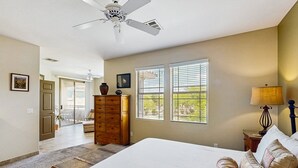 Ein Schlafzimmer mit einem großen Bett, einem Schminktisch, zwei Fenstern mit Jalousien, einem Deckenventilator und Blick ins Freie.