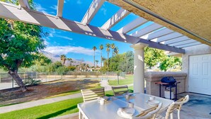Eine Terrasse im Freien mit Tisch und Stühlen, einem Grill und Blick auf einen angelegten Garten mit Palmen und einem Berg im Hintergrund.