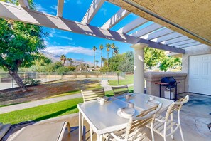 Eine Terrasse im Freien mit Tisch und Stühlen, einem Grill und Blick auf einen angelegten Garten mit Palmen und einem Berg im Hintergrund.