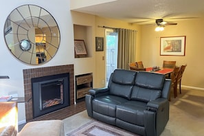 Un salon avec une cheminée, un canapé en cuir noir, une table à manger avec des chaises et un ventilateur de plafond.