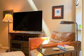 A living room with a leather sofa, a television on a wooden stand, a table with books, a lamp, and a framed picture on the wall.