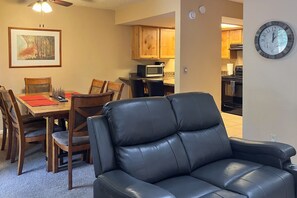 A living room with a black leather sofa, wooden dining table with chairs, a framed painting, and a kitchen in the background. - Condo with full kitchen, balconies, views of Mt. Elden - close to town (Flagstaff)