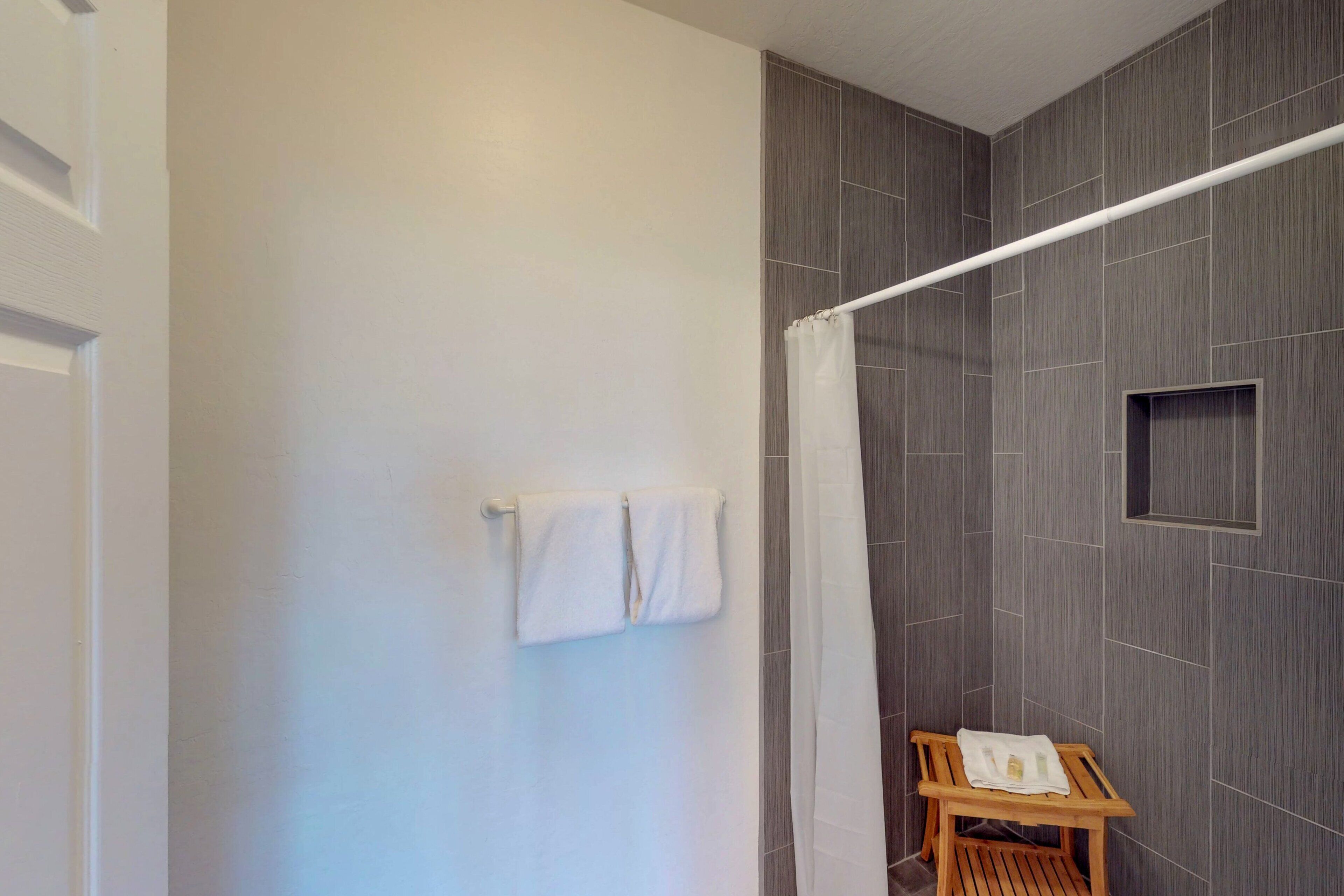A bathroom with a shower, a towel rack with two towels, and a wooden chair with a towel and a bottle on it.