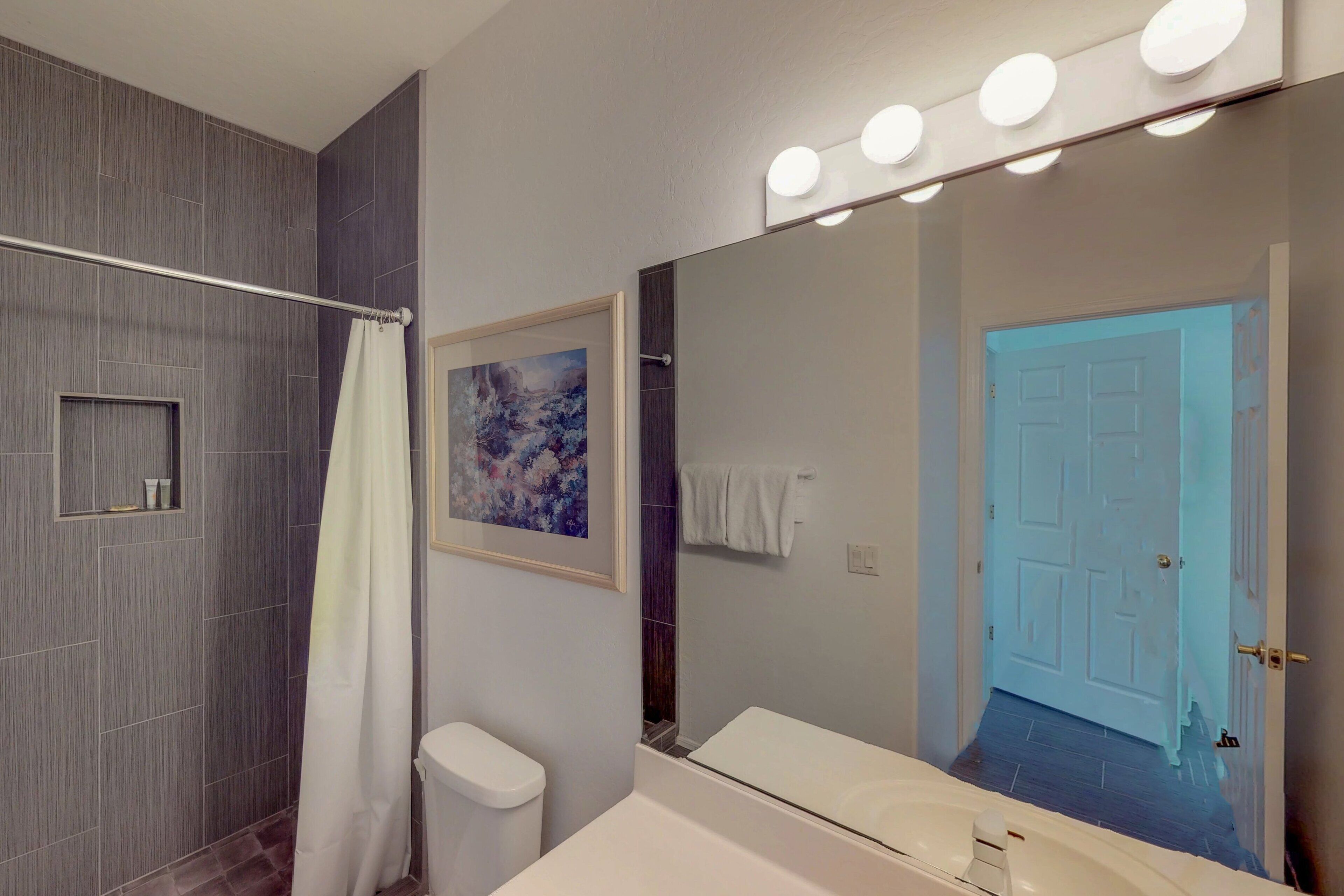 A modern bathroom with a shower, toilet, and bathtub. There is a large mirror with lights above it and a door leading to another room.