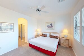 Ein Schlafzimmer mit einem Bett, zwei Nachttischen, einem Deckenventilator, einem Fenster mit Jalousien und einer Tür zu einem anderen Raum.