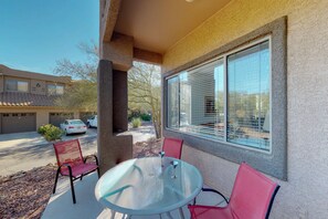 Eine Terrasse mit einem Glastisch und zwei Stühlen, ein großes Fenster mit Jalousien und ein Gebäude im Hintergrund.