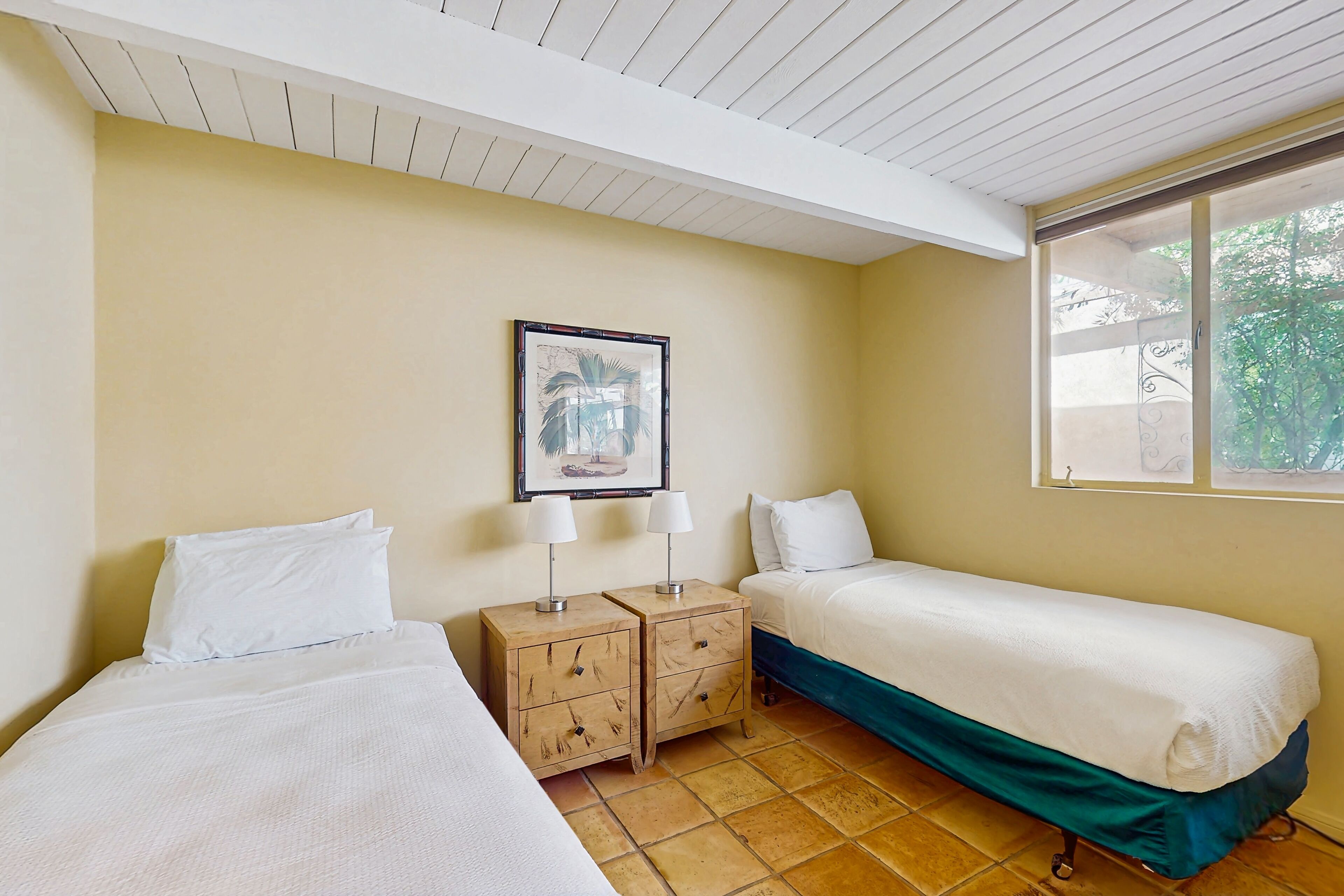 A room with two single beds, a small wooden nightstand with two lamps, a framed picture on the wall, and a window with a view of greenery.
