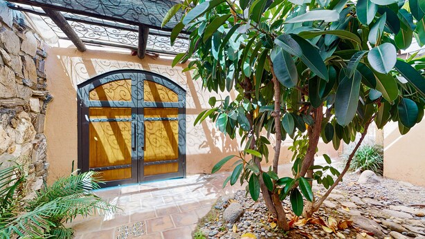 Un espacio exterior con un muro de piedra, una puerta de madera con un portón metálico y un árbol de hojas verdes.