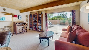 Una sala de estar acogedora con techo de madera, una lámpara roja, una estantería, un sofá de cuero y vistas a un patio exterior a través de un ventanal.