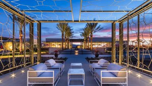 A covered outdoor seating area with lounge chairs and tables, palm trees, and a view of a building in the background. - Chic dog-friendly home with private pool, pool spa, gym, pools, grills & firepit (Palm Springs)