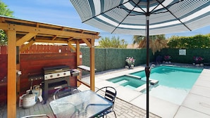 Un patio trasero con una parrilla, una mesa con sillas, una piscina y una pérgola con un parasol.