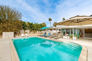 A swimming pool with lounge chairs and umbrellas, surrounded by a patio area and palm trees. - Modern home w/mountain views, private backyard pool & grill, close to downtown (Palm Springs)