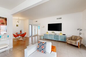 A modern living room with a white sofa, a plaid sofa, a dining area with red chairs, and a TV mounted on the wall. - Modern home w/mountain views, private backyard pool & grill, close to downtown (Palm Springs)