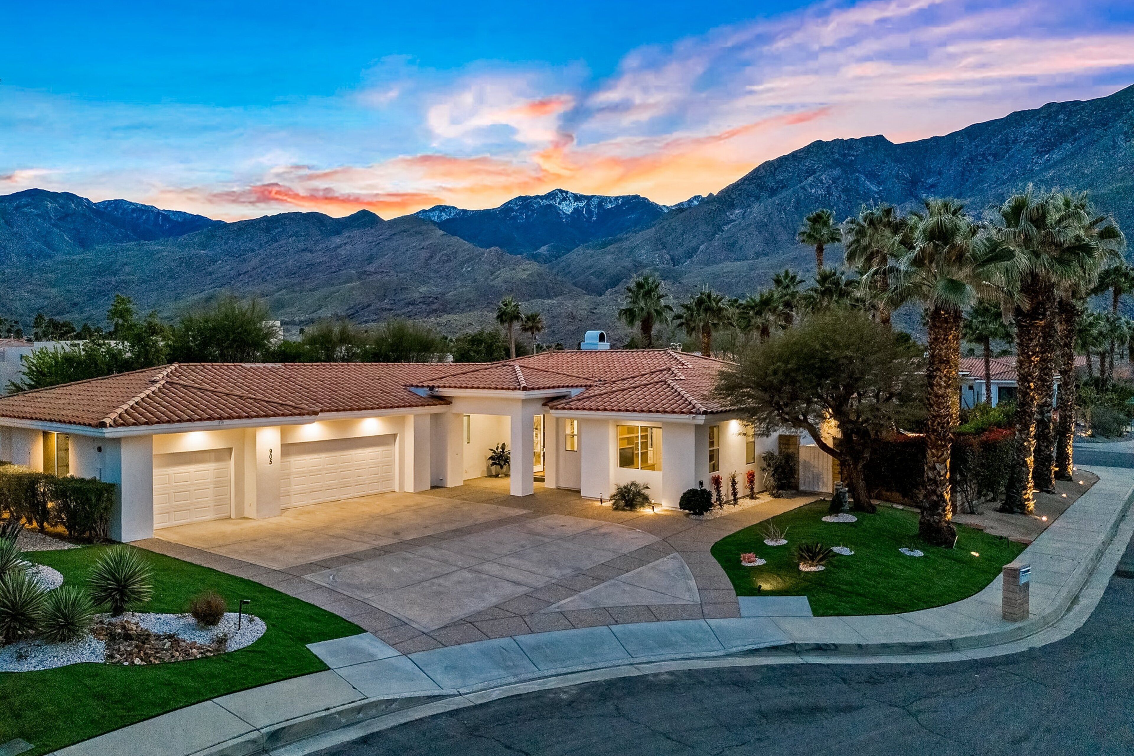 A two-story house with a garage, surrounded by a well-maintained lawn and palm trees, set against a mountainous backdrop.
