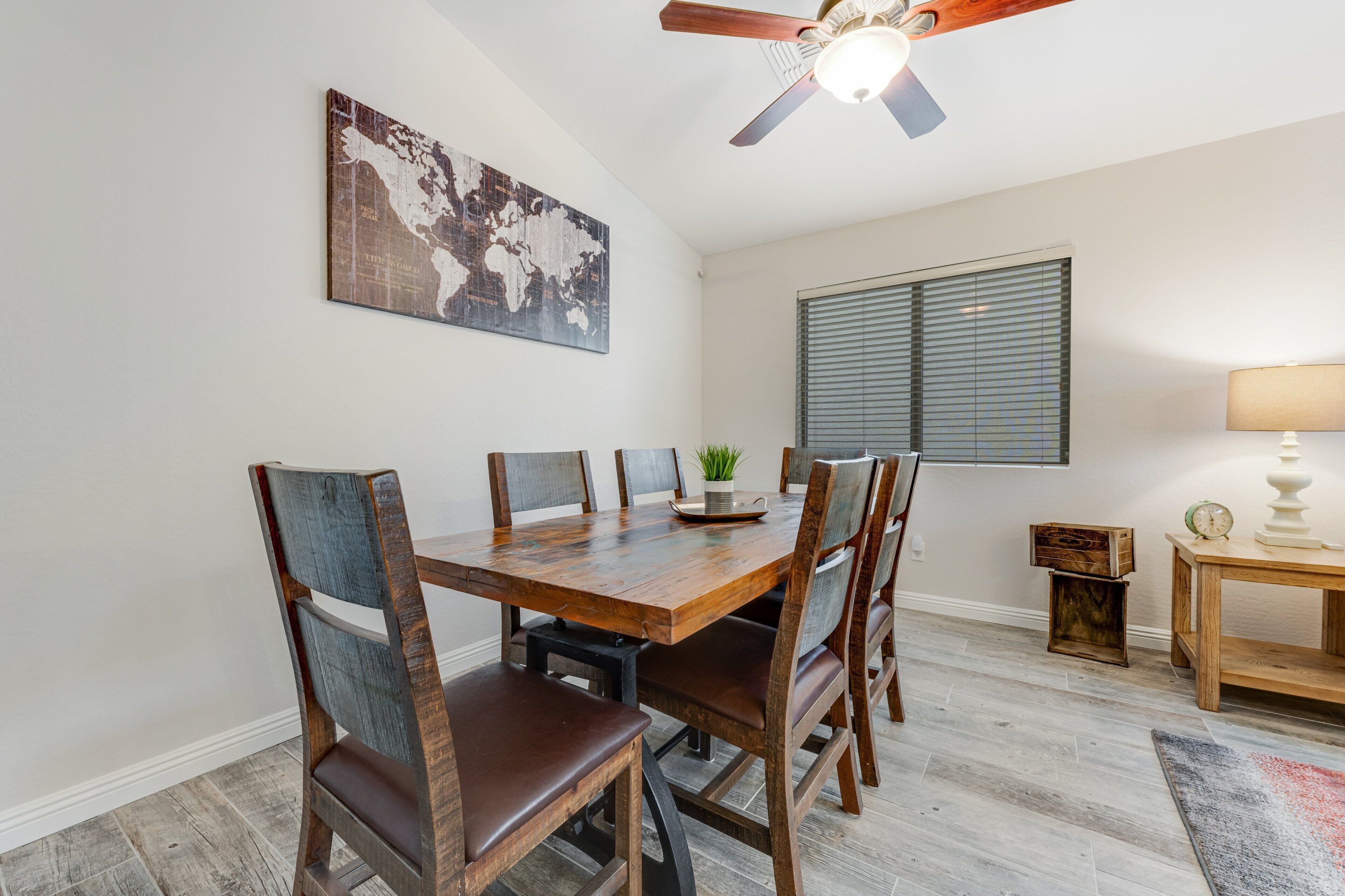 A dining area with a wooden table and chairs, a ceiling fan, a world map artwork, a window with blinds, and a small table with a lamp and clock.